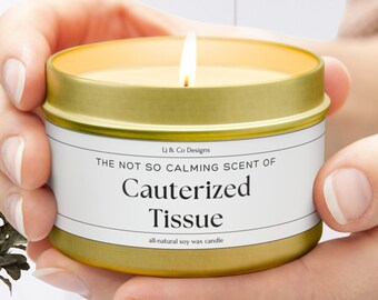 May include: A gold tin candle with a white label that reads "&Co Designs THE NOT SO CALMING SCENT OF Cauterized Tissue all-natural soy wax candle"