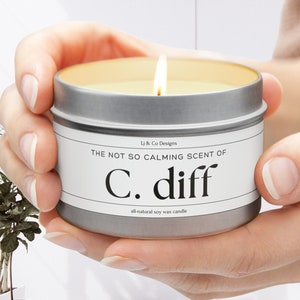 Puede incluir: Una lata de vela plateada con una etiqueta blanca que dice "The not so calming scent of C. diff all natural soy wax candle".