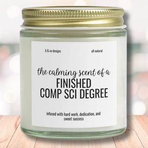 Puede incluir: Un tarro de vela blanco con tapa dorada. La etiqueta dice "the calming scent of a FINISHED COMP SCI DEGREE infused with hard work, dedication, and sweet success".