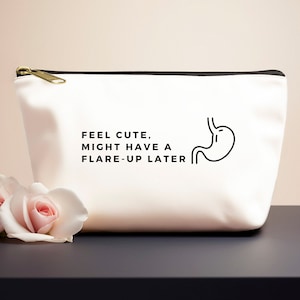 Op de afbeelding: Witte canvas toilettas met een zwarte rits en zwarte bies. De tas heeft een zwarte lijntekening van een maag met de tekst "Feel cute, might have a flare-up later".