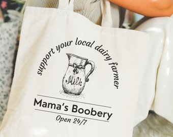 May include: A cream-coloured tote bag featuring a black illustration of a milk jug and text. The text includes "support your local dairy farmer", "Mama's Boobery", and "Open 24/7". The bag has a simple design with a handle.