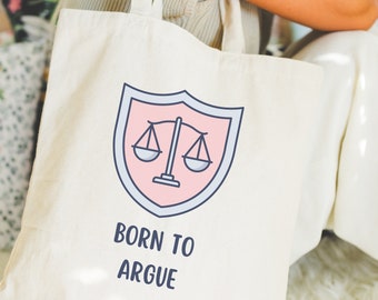 May include: A white canvas tote bag with a pink and blue illustration of a scale of justice inside a shield. The text "Born to Argue" is printed below the illustration.