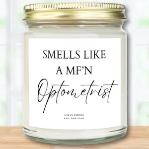 May include: A white candle jar with a gold lid and a white label that reads "Smells Like A MF'N Optometrist" with the text "LJ & CO DESIGNS 9 OZ | NON-TOXIC" at the bottom.