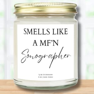 Könnte beinhalten: Ein weißes Kerzengefäß mit goldenem Deckel und einem weißen Etikett mit der Aufschrift "SMELLS LIKE A MF'N Sonographer" und "LJ & CO DESIGNS 9 OZ | NON-TOXIC".