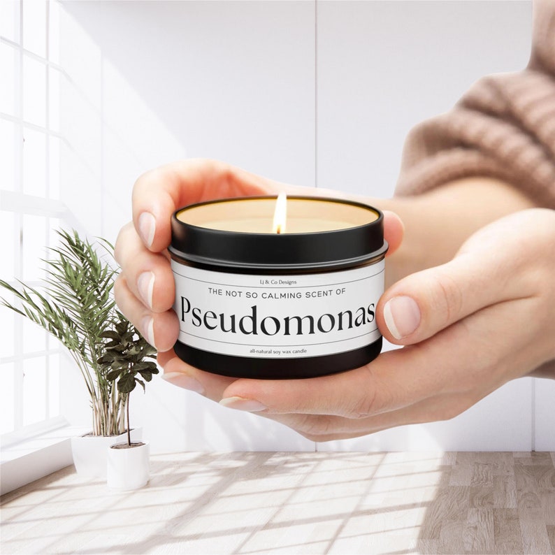 May include: A black candle jar with a white label that reads "The not so calming scent of Pseudomonas, all natural soy wax candle". The candle is lit and the flame is visible.
