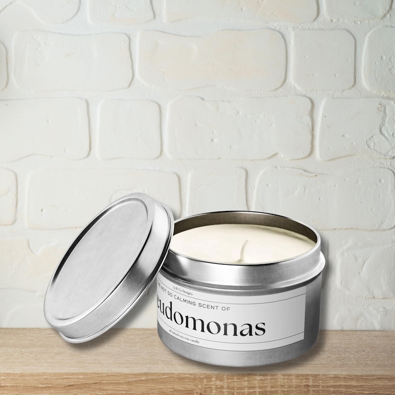 May include: A silver candle tin with a white lid. The candle is labeled "SO CALMING SCENT OF Pseudomonas".