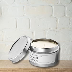 May include: A silver candle tin with a white label that reads "The Not So Calming Scent Of Bacterial Vaginosis". The candle is in a white brick background.