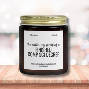 May include: A brown glass candle jar with a gold lid. The label reads "the calming scent of a FINISHED COMP SCI DEGREE infused with hard work, dedication, and sweet success".