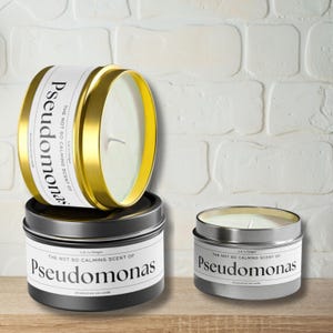 May include: Three candles in different sizes with gold and silver lids. The labels on the candles read "The not so calming scent of Pseudomonas".