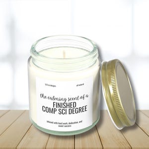 May include: A clear glass candle jar with a gold lid. The label reads "the calming scent of a FINISHED COMP SCI DEGREE infused with hard work, dedication, and sweet success".