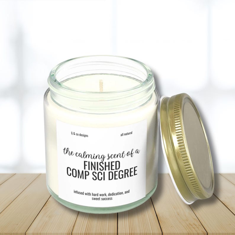 May include: A clear glass candle jar with a gold lid. The label reads "the calming scent of a FINISHED COMP SCI DEGREE infused with hard work, dedication, and sweet success".