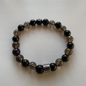 以下が含まれることがあります： 黒と灰色のビーズブレスレットに銀のチャームが付いている。ブレスレットには、黒い邪眼ビーズとクリアクォーツビーズが使用されている。