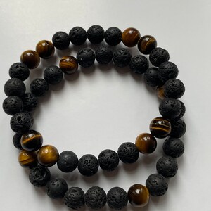 May include: Two beaded bracelets. One bracelet features black lava rock beads, and the other has a combination of black lava rock beads and tiger's eye beads. The tiger's eye beads are brown with golden stripes.