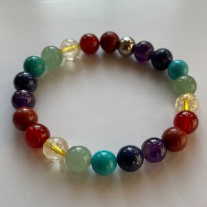 May include: A colourful beaded bracelet featuring seven different coloured beads, including red, orange, yellow, green, blue, indigo, and violet. The bracelet is made from natural stones and is designed to represent the seven chakras.