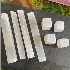 May include: Five selenite sticks and four selenite cubes on a black slate surface. The sticks are smooth and white with a striated texture. The cubes are also smooth and white.