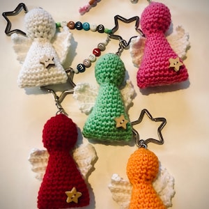 May include: Five crocheted angel keychains with white wings and star-shaped buttons. The angels are in different colors: white, pink, green, red, and orange. The keychains are connected by a silver chain with star-shaped charms.