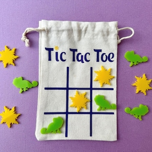 May include: A canvas bag with the words "Tic Tac Toe" printed on it, with a grid for the game. The bag is surrounded by yellow sun-shaped and green lizard-shaped game pieces, all on a purple background.