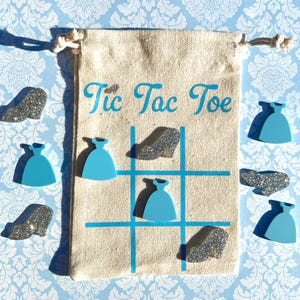 May include: A canvas Tic Tac Toe game set with a drawstring closure. The game board is printed with a blue grid and the words "Tic Tac Toe". The game pieces are blue dress shapes and silver glitter high heels.