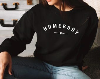 Puede incluir: Sudadera negra con el texto "HOMEBODY" en letras blancas con un pequeño corazón sobre una línea. La sudadera la lleva puesta una persona sentada en un suelo con una superficie marrón claro.