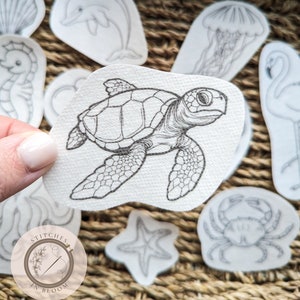 Puede incluir: Un conjunto de parches de tela blanca con ilustraciones dibujadas a mano de criaturas marinas, incluyendo una tortuga marina, un delfín, una medusa y un cangrejo. Los parches están dispuestos sobre una superficie tejida. El logotipo "Stitches in Bloom" es visible.