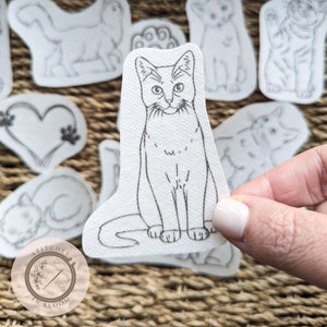 May include: A black and white line drawing of a cat sitting on a white background. The cat is looking directly at the viewer. The image is part of a set of iron-on transfers for crafting.