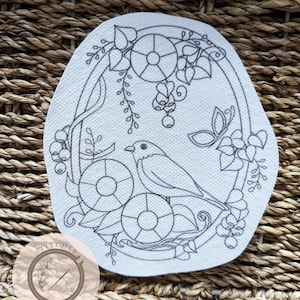 May include: A black and white line drawing of a bird sitting on a branch with flowers and berries. The drawing is on a white background and is surrounded by a circle. The text "Stitches in Bloom" is in a circle below the drawing.