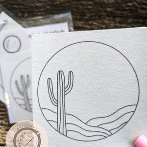 May include: A white embroidery pattern with a black line drawing of a cactus in a desert landscape. The pattern is in a circle and has the text "Stitches in Bloom" in a circle at the bottom.