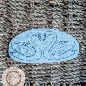 May include: A light blue patch featuring two swans facing each other, forming a heart shape with their necks. The swans are embroidered in black thread. The patch is oval-shaped and set against a woven background. A circular logo with text is in the lower left corner.