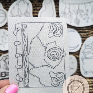 May include: A black and white line drawing of a book with a snake wrapped around it. The book has a stitched leather cover with an eye on the front. The image is on a white background with a watermark that says "Stitches in Bloom".