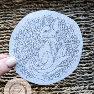 Könnte beinhalten: Ein weißer Stickrahmen mit einer schwarzen Linienzeichnung eines Fuchses, umgeben von Blumen. Der Rahmen ist mit "Stitches in Bloom" beschriftet.