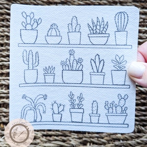 Op de afbeelding: Een witte vierkante stof met een handgestikt ontwerp van verschillende cactussen en vetplanten in potten, gerangschikt op planken. Het ontwerp is in zwart garen. In de linkerbenedenhoek staat een cirkelvormig logo met de tekst "Stitches in Bloom".