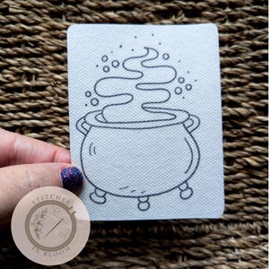 May include: A black and white line drawing of a cauldron with steam rising from it. The cauldron is on three legs and has a handle on the side. The drawing is on a white background.