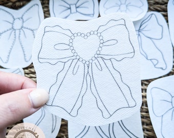 May include: A set of eight white fabric bow shapes with black outlines. The bows are various sizes and styles, including a large bow with a heart-shaped center of pearls. The fabric has a textured surface. The text "Stitches in Bloom" is visible in the lower left corner of the image.