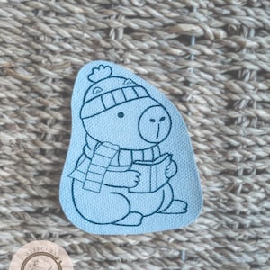 May include: A light blue fabric patch depicting a cartoon capybara wearing a hat and scarf, reading a book. The capybara is outlined in black. The patch is set against a woven background. The patch has the text "Stitches in Bloom".