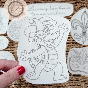 Peut inclure: Un ensemble de patchs en tissu blanc avec des motifs brodés. Un patch représente un alligator de dessin animé portant un chapeau de bouffon et un masque. D'autres patchs incluent une fleur de lys, un masque et la phrase "Laissez les bons temps rouler."