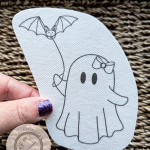 May include: A white ghost with a pink bow holding a black bat on a string. The image is on a white fabric background with the text "Stitches in Bloom" in a circle.