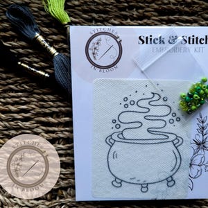 May include: A Stick & Stitch embroidery kit featuring a cauldron with green steam rising from it. The kit includes green beads and a white fabric with a black outline of the cauldron.