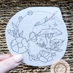 Op de afbeelding: Een wit, druppelvormig borduurpatroon met een halve maan ontwerp. Het ontwerp bevat bloemen, kristallen, paddenstoelen en sterren, allemaal zwart omlijnd. De afbeelding bevat ook het logo "Stitches in Bloom".