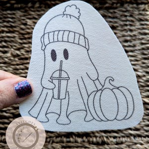 May include: A white felt appliqué of a ghost wearing a black and white striped winter hat with a pom pom on top. The ghost is holding a black and white cup with a straw and is sitting next to two black and white pumpkins. The appliqué is on a white background with the text "Stitches in Bloom" in a circle below.