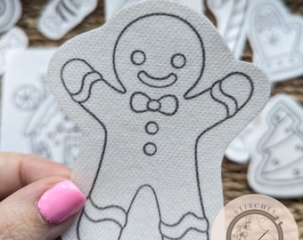 May include: A white gingerbread man-shaped felt cutout with black outline details, held in a hand with pink nail polish. Other Christmas-themed felt cutouts are in the background. The image includes a logo that says "Stitches in Bloom."