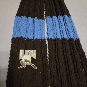 May include: A chunky knit scarf in black with two blue stripes. An embroidered patch with the word "ALASKA" and a moose silhouette is sewn on the scarf. The scarf is made of a soft, textured yarn.