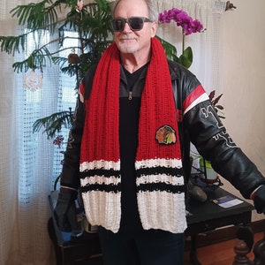 May include: A chunky knit scarf featuring horizontal stripes in red, white, and black, adorned with a circular Chicago Blackhawks team logo patch on the lower section. The scarf is worn over a black leather jacket with white and red sleeve accents.