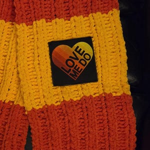 Op de afbeelding: Een dikke gebreide sjaal in oranje en geel. De sjaal heeft een vierkante patch met een hartontwerp en de tekst "LOVE ME DO". De sjaal is gemaakt van een zacht, getextureerd materiaal.