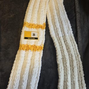 May include: A white, ribbed, chunky knit scarf featuring two horizontal yellow stripes. A rectangular patch is attached to the scarf, displaying the Vatican flag design with the word VATICAN printed in black capital letters at the bottom.