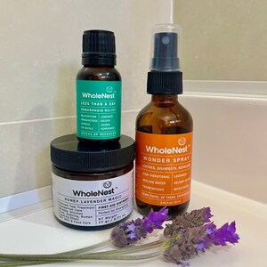 May include: Three glass bottles with black lids and labels. The bottles contain essential oils and a spray. The labels are green and orange with the text "WholeNest" and product descriptions. The bottles are on a white surface with lavender flowers.