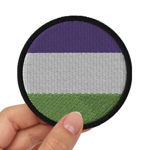 May include: A round, purple, white, and green embroidered patch with a black border. The patch is being held by a hand.