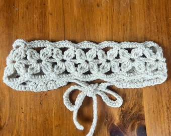 May include: A crocheted headband with a delicate floral pattern in a light beige color. The headband has a tie closure in the back.