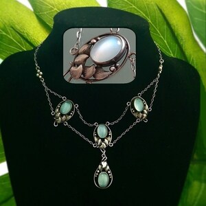 Puede incluir: Un collar de plata con un colgante de piedra lunar y piedras verdes. El colgante est&aacute; rodeado por un dise&ntilde;o floral de plata.
