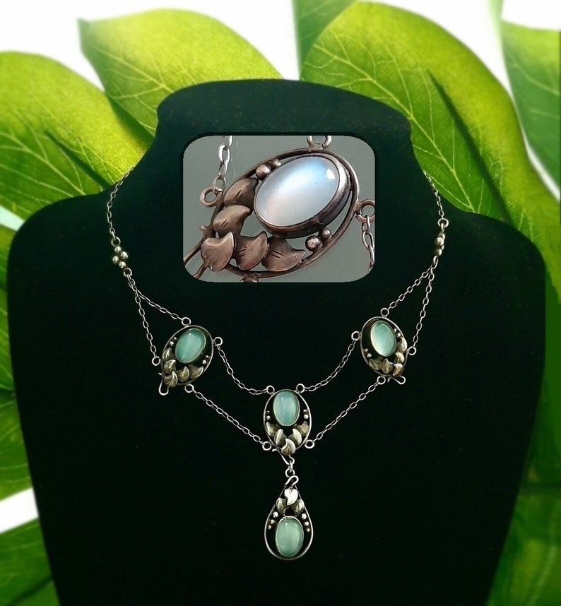 Puede incluir: Un collar de plata con un colgante de piedra lunar y piedras verdes. El colgante est&aacute; rodeado por un dise&ntilde;o floral de plata.