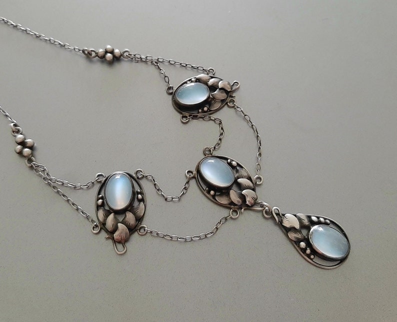 Puede incluir: Un collar de plata con una cadena delicada y cuatro colgantes ovalados de piedra lunar. Cada colgante est&aacute; engastado en un marco de plata con un dise&ntilde;o floral.